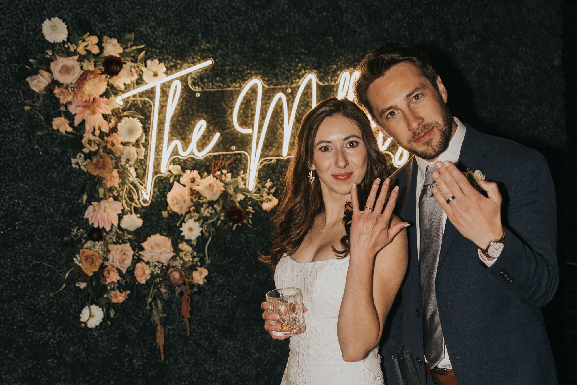 Bride and groom with neon and floral sign against green backdrop at Juniper Lodge & Treehouses in Evergree, Colorado
